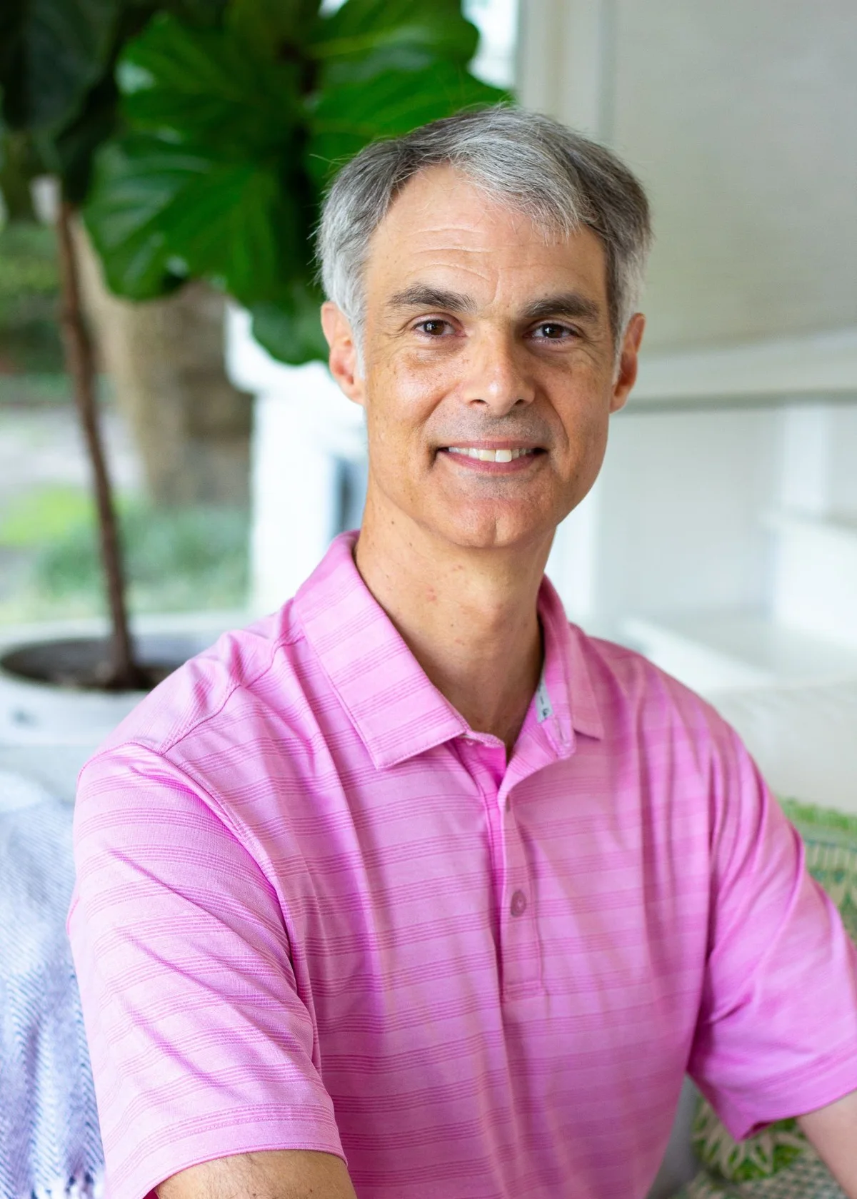 Matthew Hendley smiling at the camera in a pink polo shirt, seated by a window with a fiddle-leaf fig behind him