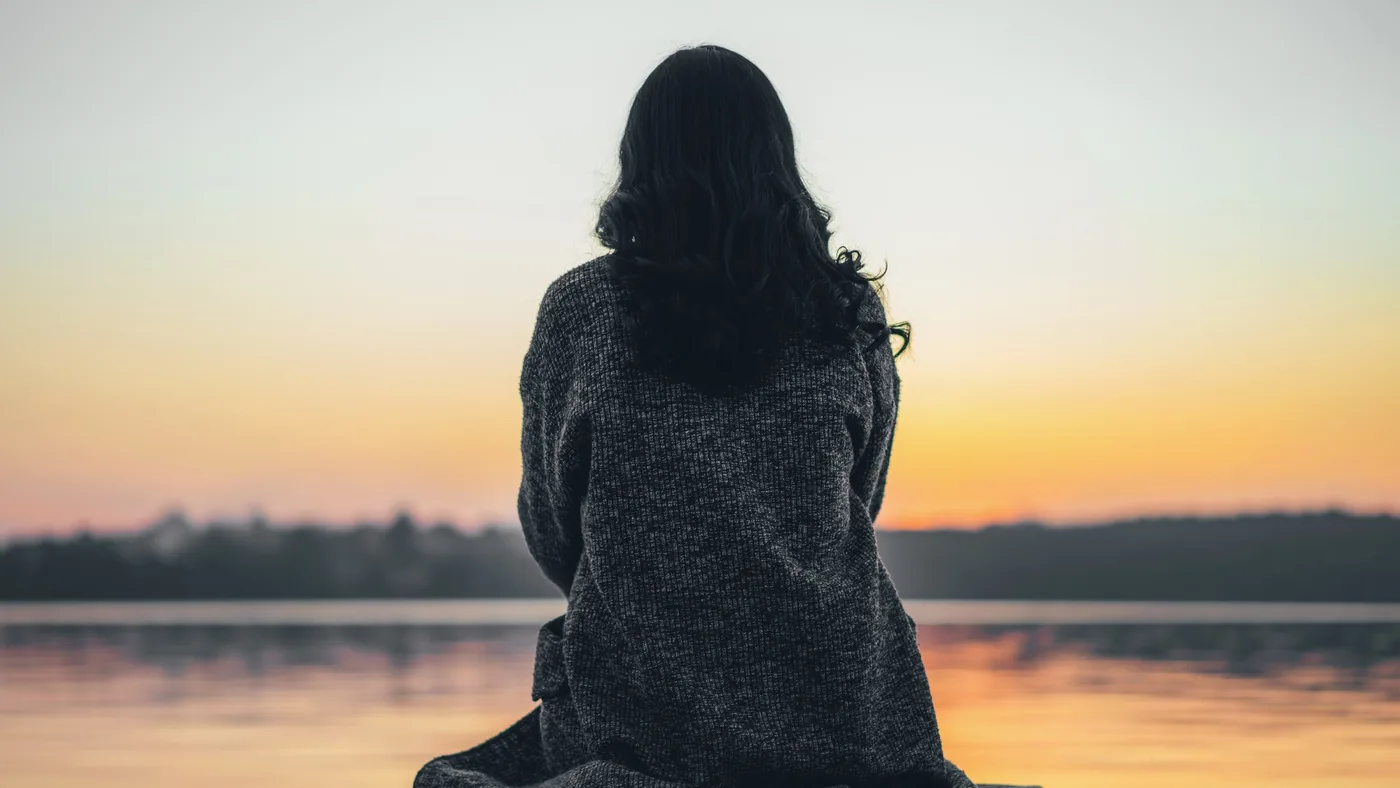 A woman wrapped in a blanket sits at the end of a dock, watching the sun set over calm water